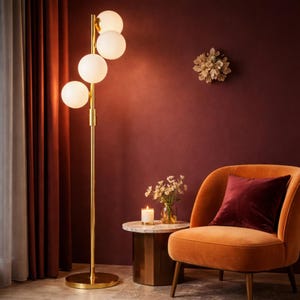May include: A modern floor lamp with a gold finish and four spherical, frosted glass shades. The lamp stands tall next to an orange armchair with a burgundy pillow. A small side table with a marble top and a candle are also visible.