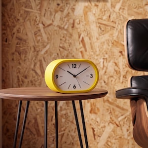 Könnte beinhalten: Eine ovale analoge Tischuhr mit einem leuchtend gelben Rahmen und einem weißen Zifferblatt. Die Uhr verfügt über schwarze Stunden-, Minuten- und Sekundenzeiger sowie schwarze Ziffern an den Positionen 12, 3, 6 und 9 Uhr und einfache Striche.
