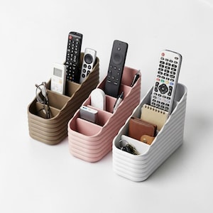 Puede incluir: Tres organizadores de escritorio de plástico acanalado en marrón, rosa y blanco. Cada organizador tiene cuatro compartimentos y contiene varios artículos, incluyendo mandos a distancia, gafas, bolígrafos y pequeños cuadernos. Sobre fondo blanco.