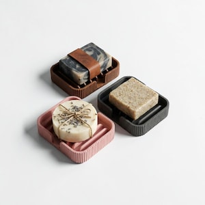 Puede incluir: Tres jaboneras en marrón, rosa y gris oscuro, cada una con una pastilla de jabón. La jabonera marrón tiene una correa de cuero alrededor del jabón. La jabonera rosa contiene un jabón redondo atado con hilo.
