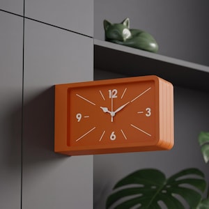 Puede incluir: Un reloj de pared rectangular con un marco naranja mate y una esfera naranja a juego. El reloj presenta marcadores de hora blancos, manecillas blancas y números blancos en las posiciones de las 12, 3, 6 y 9. El diseño es limpio y funcional.