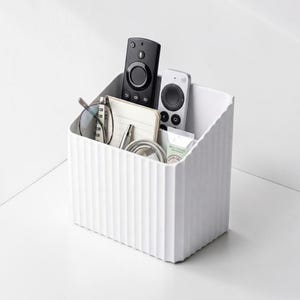 Op de afbeelding: Een witte, getextureerde, rechthoekige opbergbak met diverse items. Binnenin bevinden zich twee afstandsbedieningen, een bril, een notitieblok, een pen, een kabel en een kleine tube crème. De bak staat op een wit oppervlak.