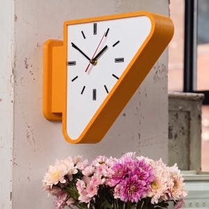 Puede incluir: Un reloj de pared con un marco triangular de color naranja brillante y una esfera blanca. El reloj presenta marcadores de hora negros, manecillas de hora y minutos negras y un segundero rojo fino. Diseño geométrico con soporte lateral.