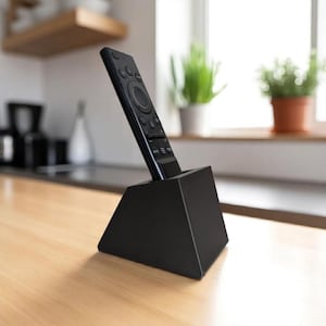 Op de afbeelding: Een zwarte afstandsbediening is geplaatst in een matzwarte, wigvormige standaard. De afstandsbediening is voorzien van een cirkelvormig navigatiepad, diverse functieknoppen en een numeriek toetsenbord. De standaard heeft een brede basis.