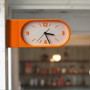 Puede incluir: Un reloj de pared de doble cara con un marco naranja oblongo y una esfera blanca. El reloj presenta números naranjas en las posiciones de las 3, 6, 9 y 12, marcas naranjas para las otras horas y manecillas negras para horas, minutos y segundos.
