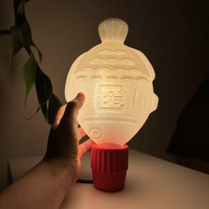 May include: A unique, glowing lamp with a fish-shaped shade. The shade is a translucent off-white color, and the base is a bright red. The lamp has a textured surface and a small, round base. The lamp is plugged into a black cord.