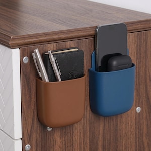 Op de afbeelding: Twee aan de muur gemonteerde opbergcontainers, één bruin en één blauw, bevestigd aan een houten oppervlak. De bruine container bevat twee pennen en een klein zwart notitieboekje. De blauwe container bevat een smartphone en een paar zwarte draadloze oordopjes.