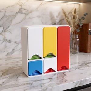 Op de afbeelding: Een witte kunststof theezakjeshouder met vijf vakken en kleurrijke voorpanelen. De bovenste rij heeft een wit, een geel en een hoog rood paneel. De onderste rij heeft een blauw en een wit paneel. In elk vak zijn theezakjes zichtbaar.