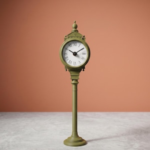 Op de afbeelding: Een groene, vrijstaande klok met een witte wijzerplaat en zwarte Romeinse cijfers. De klok is gemonteerd op een lange, slanke paal met een ronde basis. De achtergrond is effen oranje.