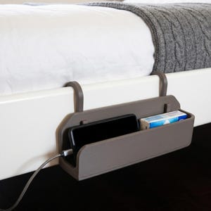 Peut inclure: Un organisateur de chevet en plastique gris mat doté de deux crochets pour se fixer au cadre d'un lit. Il contient un smartphone noir relié à un câble de charge et une petite boîte rectangulaire avec un emballage bleu et blanc.