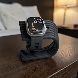 Op de afbeelding: Een donkergrijze smartwatch met een rechthoekig scherm dat de Eiffeltoren en de tijd 22:42 weergeeft, rustend op een donkergrijze, geribbelde, gebogen standaard. Het horloge heeft een gestructureerde siliconen band en een metalen behuizing met een oranje knop.