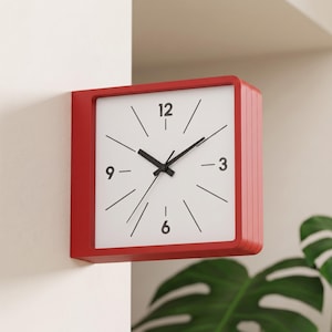 Peut inclure: Une horloge murale carrée avec un cadre rouge vif et un cadran blanc. L'horloge présente des chiffres noirs aux positions 12, 3, 6 et 9, avec de fines lignes noires marquant les autres heures. Les aiguilles noires des heures, minutes et secondes sont centrées.