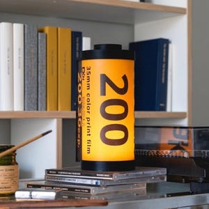 Könnte beinhalten: Eine zylindrische Lampe, die wie eine 35mm-Farbfilmrolle gestaltet ist. Der Körper ist leuchtend gelb mit großem schwarzem Text, der 200 liest, sowie kleinerem Text, der 35mm color print film, DX und 200 36exp liest. Sie hat schwarze Kappen.