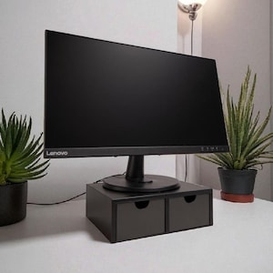 May include: A black Lenovo computer monitor on a dark grey monitor stand with two drawers. The stand is on a white desk, with potted plants either side. A wall lamp is visible in the background.