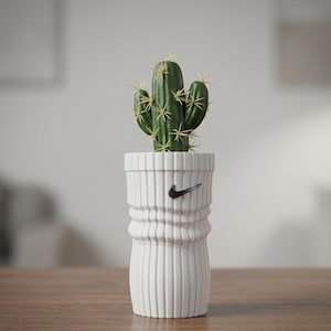Può includere: Un cactus verde con spine gialle appuntite piantato in un vaso cilindrico bianco. Il vaso presenta una texture a coste verticali e un piccolo logo Nike swoosh nero centrato sulla parte superiore della superficie esterna.