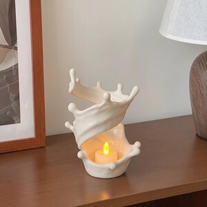 May include: A cream-colored ceramic candle holder shaped like a swirling liquid splash. The structure features an open, organic design with rounded edges and a hollow center that contains a small, battery-operated LED tea light with a warm glow.