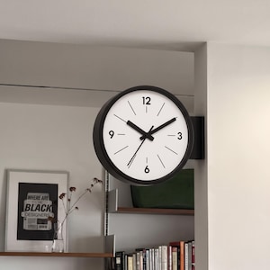 Op de afbeelding: Een dubbelzijdige wandklok met een zwarte ronde rand en een witte wijzerplaat. De klok heeft zwarte uur-, minuut- en secondewijzers, met zwarte cijfers op de posities 12, 3, 6 en 9 en eenvoudige zwarte streepjes voor de overige uren.