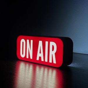 Könnte beinhalten: Ein rechteckiges, beleuchtetes "ON AIR"-Schild mit rotem Hintergrund und weißer Schrift. Das Schild hat abgerundete Ecken und einen schwarzen Rahmen. Das Bild ist vor einem dunklen Hintergrund, wobei das Schild ein rotes Leuchten wirft.