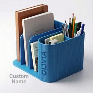 Pode incluir: Um organizador de secretária azul com vários compartimentos. Contém cadernos, livros, canetas, lápis e notas autocolantes. O organizador tem o nome "OLIVIA" na lateral e as palavras "Custom Name" por baixo.