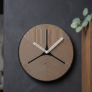 Op de afbeelding: Een ronde wandklok met een lichte houten afwerking en een reeks concentrische, U-vormige latten die een geometrisch patroon vormen. De klok heeft twee dunne, witte wijzers in het midden tegen een zwarte achtergrond.