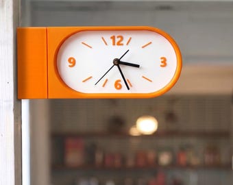 Può includere: Un orologio da parete bifacciale con una cornice rettangolare arrotondata di colore arancione brillante. Il quadrante bianco presenta numeri arancioni alle ore 3, 6, 9 e 12, tacche arancioni per le altre ore e lancette nere per ore, minuti e secondi.