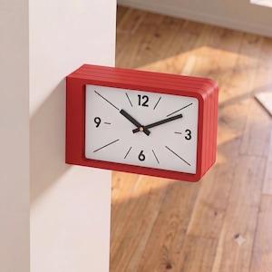 Puede incluir: Un reloj de pared rectangular con un marco rojo brillante y una esfera blanca. El reloj presenta marcadores de hora negros, manecillas negras y los números 12, 3, 6 y 9 impresos en una fuente negra negrita. El reloj está montado en una pared.