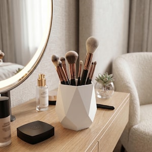 Puede incluir: Un porta brochas de maquillaje geométrico blanco que contiene varias brochas con mangos negros y virolas de color cobre. Las brochas tienen cerdas suaves y claras de varios tamaños, dispuestas verticalmente dentro del recipiente facetado.