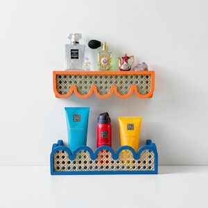 Op de afbeelding: Twee decoratieve planken, één oranje en één blauwe, met een geweven rietontwerp. De bovenste plank bevat parfumflesjes en een kleine theepot. De onderste plank bevat blauwe en gele cosmetische tubes en een rode spuitfles.
