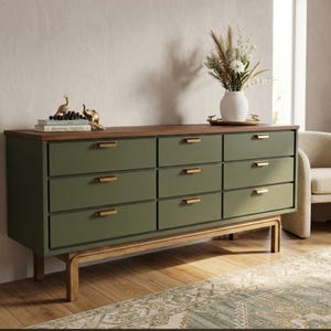 May include: A long dresser with nine olive green drawers, each featuring a slim gold handle. The unit has a dark wood top surface and is supported by a light wood frame with four legs. A white vase with dried flowers sits on the top surface.