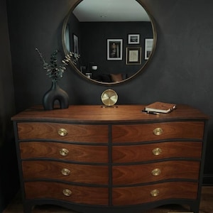 May include: A wooden chest of drawers with eight drawers featuring a dark frame and natural wood grain fronts, accented by brass hardware. On top sits a matte black vase with eucalyptus, a gold circular clock, and a leather-bound notebook with a pen.