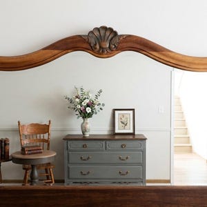 Puede incluir: Espejo de madera grande y ornamentado con un diseño de concha tallada en la parte superior. El espejo refleja una habitación con una cómoda gris, una pequeña silla de madera y un jarrón con flores. También se ve un cuadro enmarcado y una mesita.