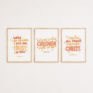 Può includere: Tre stampe da parete rettangolari in cornici di legno chiaro. Ogni stampa bianca presenta versetti biblici scritti a mano in arancione e rosso: When I am afraid I put my trust in you, Let the little children come to me, e I can do all things through Christ.