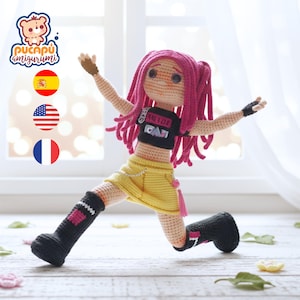 May include: A handmade crochet doll with pink hair, a black top, and a yellow skirt. The doll is wearing black boots and has its arms outstretched. The doll is in a dynamic pose, as if running or dancing. The doll has a logo in the top left corner.