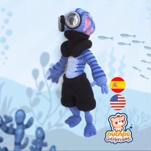 Puede incluir: Muñeco amigurumi hecho a mano que representa a un buzo, con casco de buceo, pantalones cortos negros y una bufanda negra. El cuerpo es azul y blanco a rayas. El fondo es azul claro, con ilustraciones de peces.