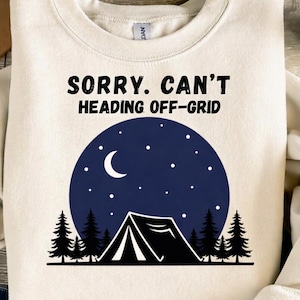 May include: Cream-colored sweatshirt with the text "SORRY. CAN'T HEADING OFF-GRID" in black. Below the text is a dark blue circle with a crescent moon and stars, and a black tent and trees.