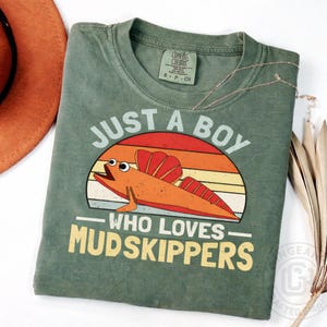 May include: Green Comfort Colors t-shirt with the text "JUST A BOY WHO LOVES MUDSKIPPERS" and a cartoon mudskipper design. The design features a sunset background with orange, yellow, and red hues.