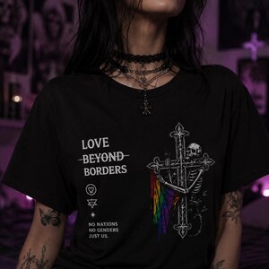 May include: A black t-shirt featuring a white graphic of a skeleton embracing a cross with a rainbow flag draped over it. The text reads LOVE BEYOND BORDERS, NO NATIONS NO GENDERS JUST US, along with small symbols including a heart and a star.