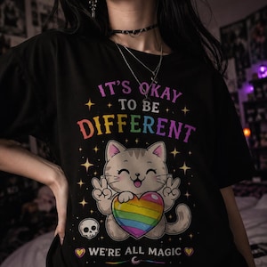 Puede incluir: Camiseta negra con el texto "IT'S OKAY TO BE DIFFERENT" en colores arcoíris. Un gato de dibujos animados sosteniendo un corazón arcoíris aparece debajo del texto. La camiseta también incluye la frase "WE'RE ALL MAGIC" y una calavera pequeña.