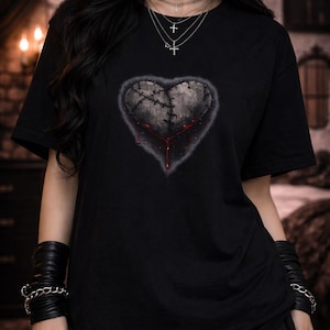 May include: Black t-shirt featuring a distressed heart design with visible stitching and dripping blood. The heart is gray with a dark outline. The model is wearing multiple necklaces and bracelets, with a black choker. The overall theme is gothic.