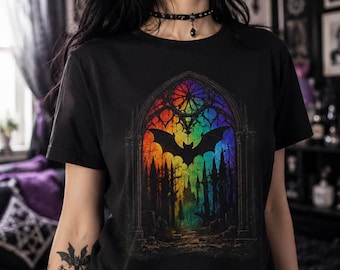 May include: A black short-sleeved t-shirt featuring a central graphic of a rainbow-colored stained glass window archway. The design depicts a silhouette of a bat flying above a dark, silhouetted forest landscape within the arched frame.