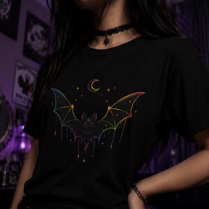 May include: A black short-sleeved t-shirt featuring a central graphic of a bat with rainbow-colored wings. The design includes a crescent moon and small stars above the bat, with colorful lines dripping downward from the wing tips.