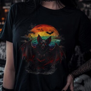 May include: A black short-sleeved t-shirt featuring a graphic print of a bat with red eyes and spread wings. The bat is positioned in front of a circular background with horizontal stripes in orange, yellow, and green, with small bat silhouettes.