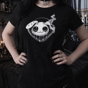Może przedstawiać: Czarny t-shirt z kreskówkowym wzorem lalki, z przeszytymi detalami, dużymi oczami i ociekającymi akcentami. Projekt jest umieszczony w okrągłej ramie. Modelka ma na sobie czarny choker z wisiorkiem.