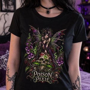 May include: A black short-sleeved t-shirt featuring a graphic print of a fairy with dark hair and wings sitting on a skull. The design includes purple foxglove flowers, green vines, and small skulls. The text "POISON PIXIE" is printed in a stylized font.