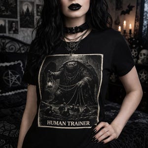May include: Black t-shirt featuring a detailed illustration of a creature holding a lantern and controlling figures with strings. The text "HUMAN TRAINER" is printed below the image. The shirt is a crew neck style.