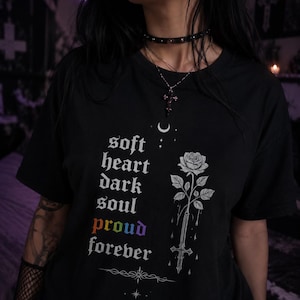 May include: Black t-shirt with the words "soft heart dark soul proud forever" in white gothic font. A white rose and sword graphic is on the right side. The shirt has short sleeves.