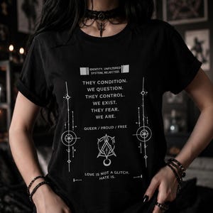 May include: Black t-shirt with white text and graphic design. The text includes phrases like "Identity, Unfiltered, System, Rejected," and "Love is not a glitch. Hate is." The shirt features geometric and symbolic designs, with the words "Queer / Proud / Free."