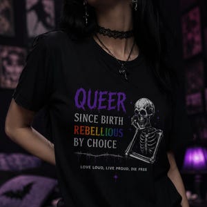 May include: A black t-shirt featuring a graphic of a skeleton resting its chin on its hand. The text reads: QUEER SINCE BIRTH, REBELLIOUS BY CHOICE, and LOVE LOUD, LIVE PROUD, DIE FREE. The word QUEER is purple, and REBELLIOUS is in rainbow colours.