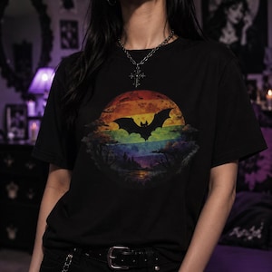May include: A black short-sleeved t-shirt featuring a circular graphic print. The design displays a silhouette of a bat flying in front of a vibrant, multi-colored sunset or moon, with dark tree silhouettes and a landscape visible at the bottom.