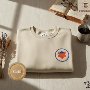 Può includere: Una felpa color sabbia con scollo tondo e una toppa ricamata circolare con una volpe arancione con un cappello blu. La toppa è bordata di blu. La parola "EMBROIDERED" è sopra la felpa.